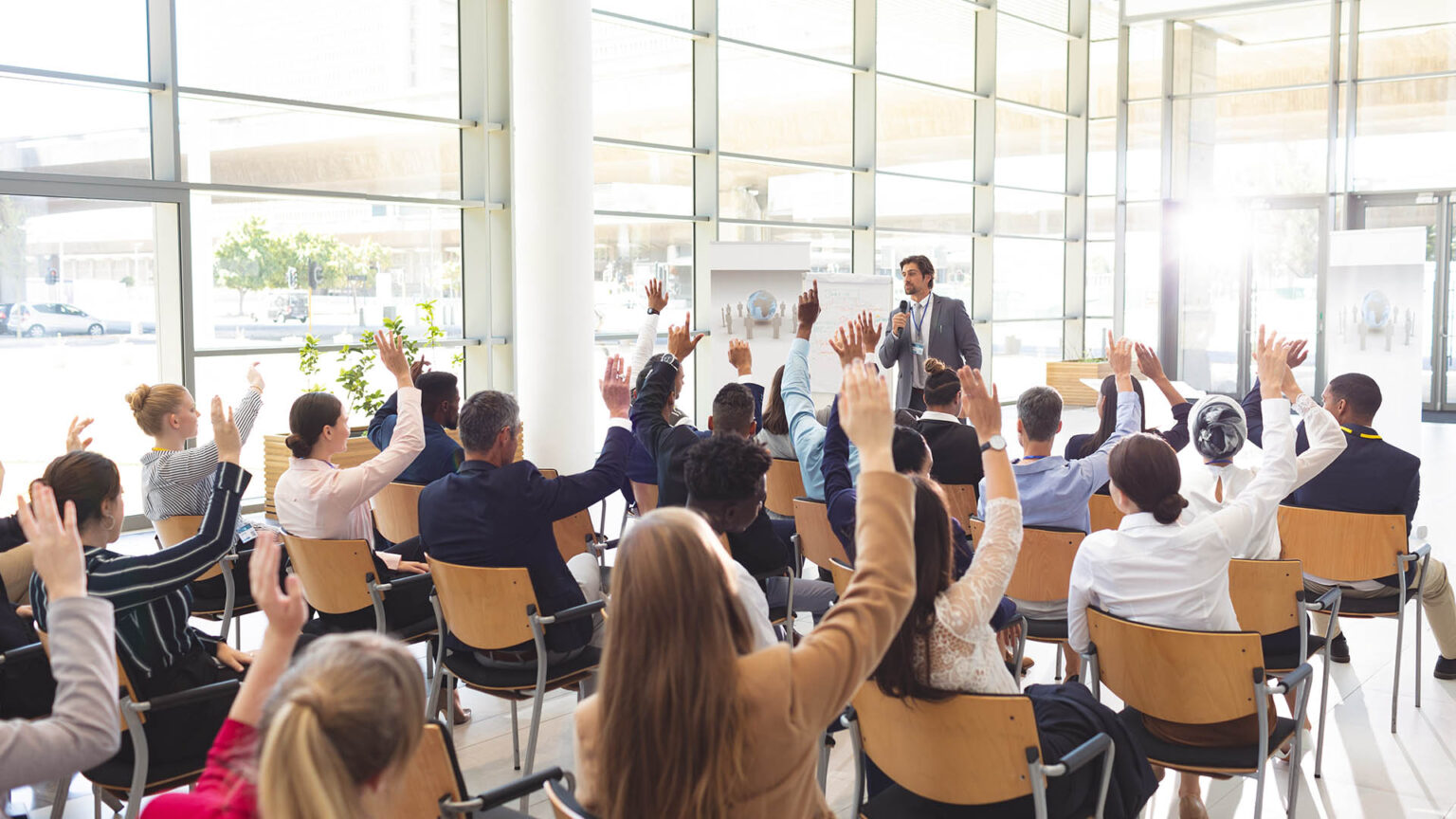 Rear view of interactive diverse business people listening to Caucasian businessman and raising hands to ask him questions in conference